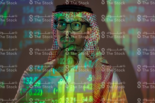 شاب سعودي يرتدي الزي السعودي التقليدي يتابع سوق الأسهم ويظهر انعكاس أضواء الشاشة عليه بينما يضع يده على دقنة يفكر, رجل يتابع سوق المال