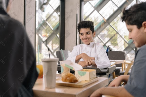 طفل "من ذوي الإعاقة" يرتدي الزي السعودي يجلس على كرسي متحرك ويتناول الطعام مع شخصين آخرين في مقهى.