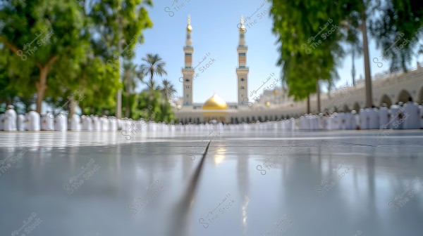 An outdoor scene featuring a mosque with two large golden domes and tall minarets in the background. In the foreground, there is a wide courtyard with polished tiles reflecting light. The area is surrounded by green trees and people dressed in traditional white clothing, appearing in rows.