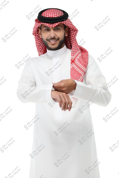Image of a man wearing traditional Saudi attire, a white thobe with a red and white checkered shemagh and a black agal. The man is smiling slightly as he adjusts his left cuff with his right hand. The background is white.