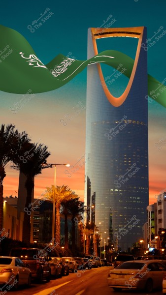 The Kingdom Tower in Riyadh, Saudi Arabia, is shown at sunset, with a sky colored in shades of orange and blue. The tower is topped with a flagpole carrying the flag of Saudi Arabia. In the foreground, a street lined with palm trees and parked cars on both sides.