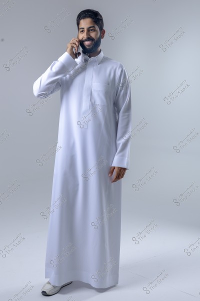 A man wearing a white thobe is talking on the phone with a smile, standing in a studio environment with a gray background. The thobe appears to be of the traditional style worn in the Gulf region. The man is wearing white shoes and seems content and engaged in the conversation.