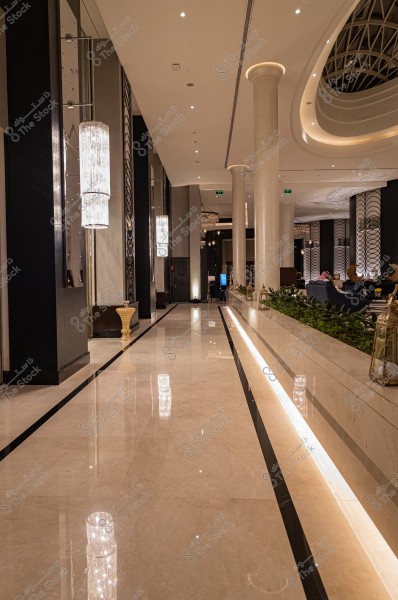 An image of a luxurious hotel lobby featuring a shiny marble floor and tall white columns. Elegant chandeliers hang from the ceiling with a series of wall lights. Modern decor adorns the walls, and there are carefully arranged seating areas in the background where some people are seated. Plants neatly decorate the sides.