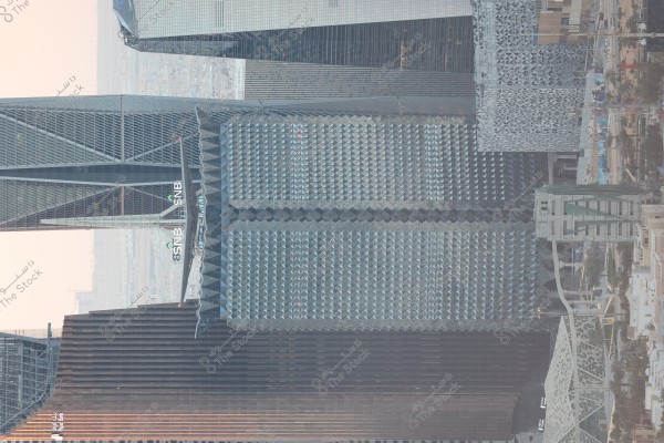 An image of modern high-rise buildings in Riyadh, Saudi Arabia. The photo highlights a notable tower with a distinctive glass design featuring geometric patterns, and the name \"SNB\" is visible on the building. Surrounding buildings exhibit contemporary architecture, with a street and some trees visible below.