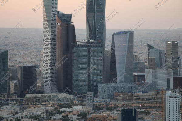 Image of the skyline of Riyadh featuring a collection of modern skyscrapers with diverse architectural designs. The scene includes prominent towers such as the Kingdom Tower and Al Faisaliah Tower, amidst other uniquely designed contemporary buildings. The background shows an expansive cityscape under a pale sky at dusk.