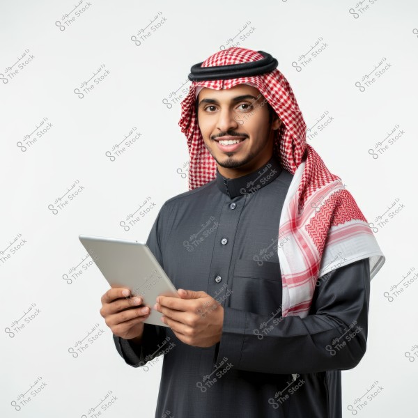 A portrait of a man wearing a dark-colored thobe and a red and white checkered headscarf with an agal. He is holding a tablet in his hands and smiling. The image clearly shows traditional Saudi attire.