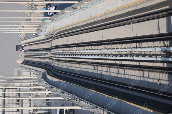 View of a modern train traveling on an elevated railway track in an urban cityscape. The train is visible in the foreground, with tracks surrounded by metal structures and pillars. In the background, city buildings and a few cars under the bridge are visible. There is a clear sky, indicating a sunny day.