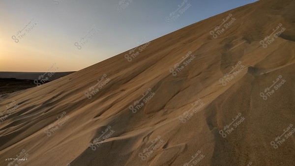 تلال رملية مرتفعة في الصحراء تحت ضوء الشمس الغاربة, تظهر السماء بخلفية زرقاء فاتحة وتميل نحو الأفق, حيث تظهر الشمس بشكل خافت في الأفق البعيد.