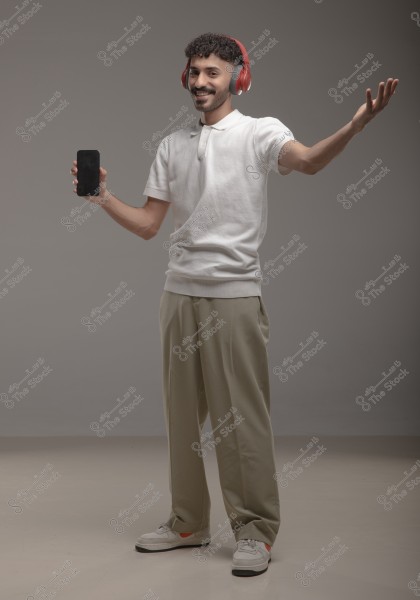 A portrait of a smiling man standing, wearing red headphones, a white shirt, and beige pants. The man holds a smartphone in one hand and extends his other arm to the side. He appears to be in a photography studio with a gray background. He is wearing white sneakers.