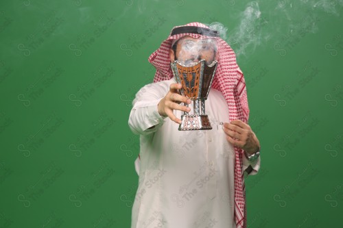 رجل سعودي يقف على خلفية خضراء يرتدي الثوب و الشماغ، يقوم بعمل حركات بيديه ، يبخر نفسه بالطيب و العود، احتفالات العيد