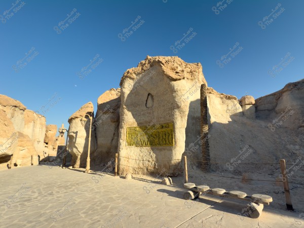 يظهر في الصورة جدار صخري كبير عليه لوح ذهبي يحتوي على نقوش بارزة. الأرضية مرصوفة بالحجارة ويبدو مصباح إنارة في الخلف على جانب الجدار. السماء زرقاء وصافية, مما يشير إلى جو مشمس ونقي.