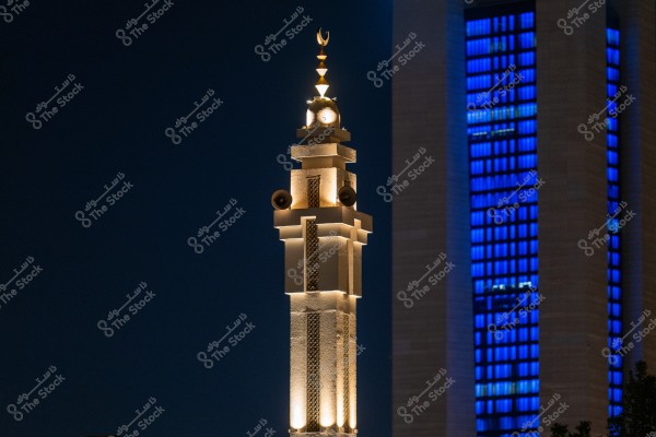 Nighttime image of a section of a mosque minaret illuminated with warm white lights, featuring a small dome and crescent at the top. Loudspeakers are visible on the sides. In the background, a building with bright blue lights in its windows can be seen, creating a contrast between the blue and white lighting.