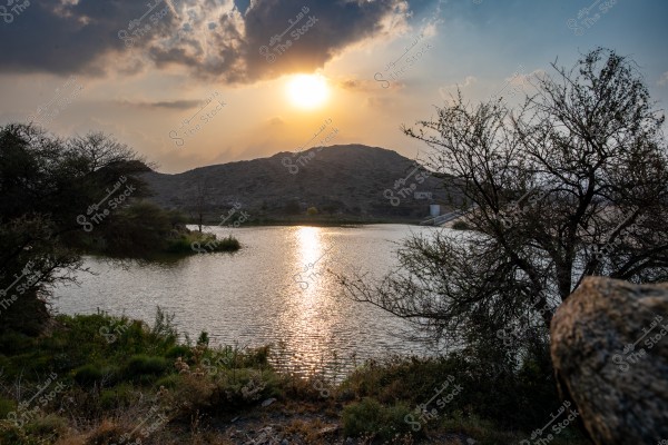 The image shows a natural landscape featuring a lake surrounded by greenery and trees. The sun is setting behind the mountains in the background, casting golden rays on the water\'s surface. The sky is filled with clouds reflecting on the lake.