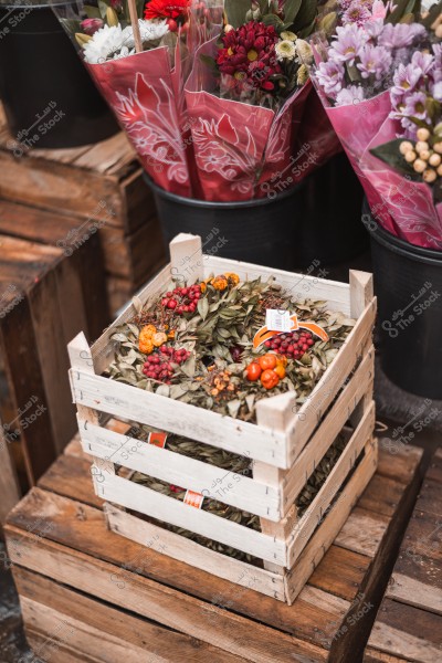 A wooden crate containing dried leaves and colorful berries, surrounded by bouquets of wrapped flowers in the background.