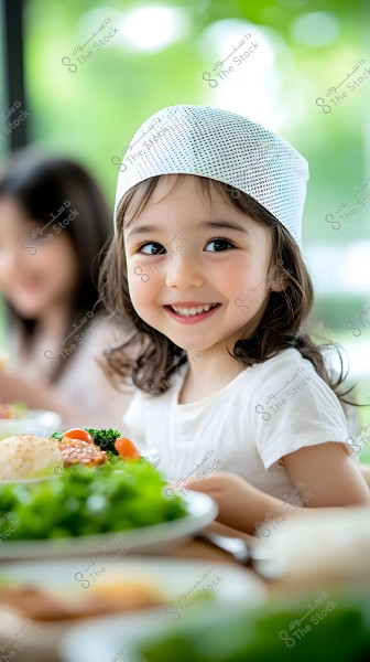 Image of a smiling young girl sitting at a table filled with healthy foods such as vegetables and salad. The girl is wearing a white shirt and a white mesh head covering, with a blurred figure of another person in the background. The environment appears bright and cheerful with a green background, possibly indicating a window view of nature.