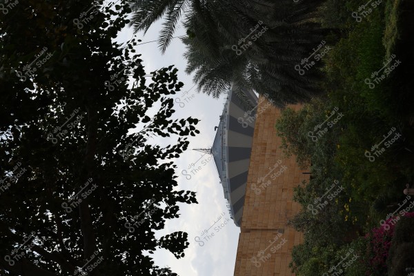 The image shows a building with a distinctive geometric design, featuring a cone-shaped roof. It is surrounded by palm trees and various plants, including dense-leaved trees and flowering shrubs with multiple colors. The sky is overcast in the background, adding to the overall atmosphere of the image.