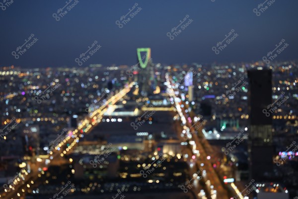 A blurred night-time photo of a city that appears to be in Saudi Arabia, with vibrant city lights and a brightly lit street stretching into the horizon. The iconic Kingdom Tower stands in the middle with its distinct architecture illuminated in green and yellow.