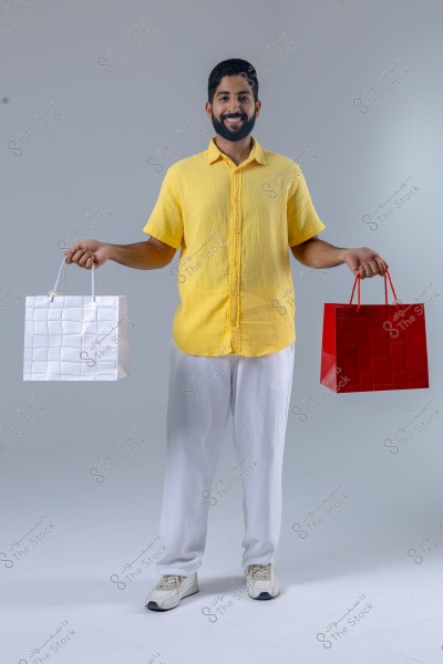The image shows a man standing wearing a yellow shirt, white trousers, and white sneakers, holding two shopping bags, one white and the other red. The background is plain gray, emphasizing the man\'s figure and the bags he is holding.