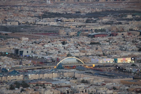 صورة جوية لمدينة الرياض في السعودية, تظهر عددًا كبيرًا من المباني السكنية الحديثة والمجمعات التجارية. في الوسط, يظهر جسر ذو تصميم حديث بقوس مضيء وعليه زجاج. في الخلفية, تتنوع المباني بين مراكز تسوق ومباني مرتفعة وأراضٍ مفتوحة.