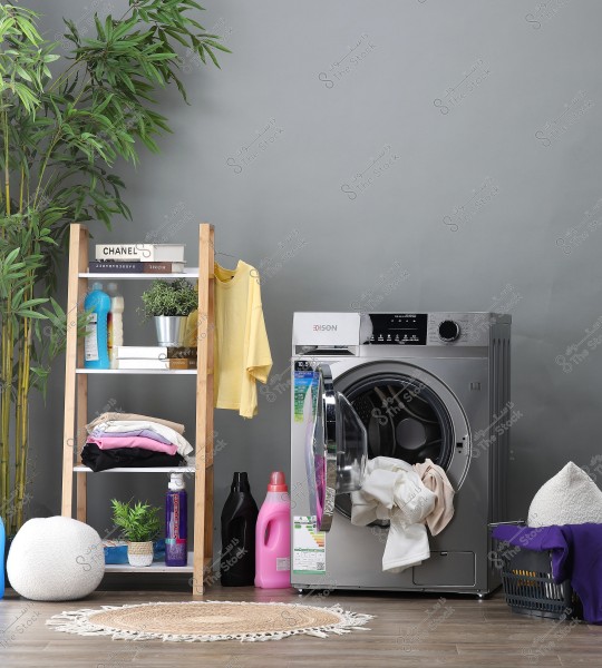 A stylish laundry room features a modern silver washing machine with its door open, containing some white clothes. Next to the washer, there is a plastic laundry basket filled with clothes. To the left, there\'s a wooden shelf with colorful detergents, books, small decorative plants, and a hanging yellow shirt. In the foreground, there is a small round rug and a large potted plant atop the shelf. The background is gray.