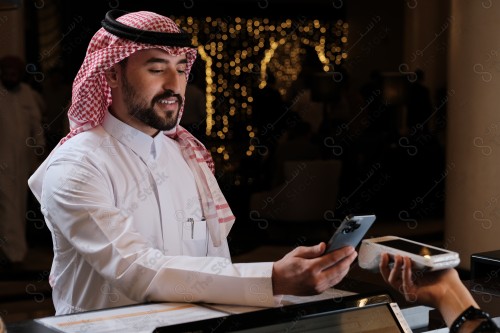 شاب سعودي في استقبال الفندق ، ويقوم بتعابير مختلفة باستخدام الجوال  ، ويقوم بدفع عبر جهاز الدفع الالكتروني