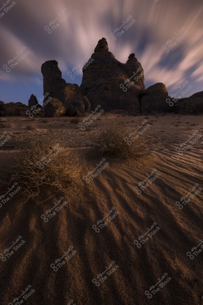 A landscape image showing large rock formations under warm daylight. The sky is filled with moving clouds, giving a sense of motion. In the foreground, there are rippled sands and scattered desert plants casting shadows on the ground.