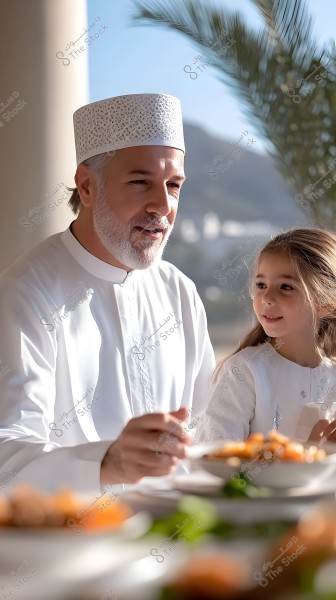 A man wearing a white robe and a traditional cap is sitting next to a young girl in a white dress at a dining table with various dishes. The background features a natural scene with palm trees and a mountain in the distance. The traditional clothing suggests they may be in a Gulf country or Oman.