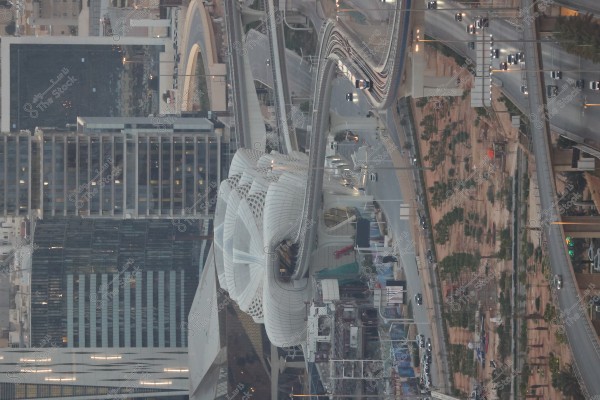 An image of an urban cityscape featuring modern skyscrapers and a metro line passing through a wide bridge. The architecture includes an innovative white structure prominently in the center. The right side of the image shows a busy highway with cars, while on the left, there are tall buildings.