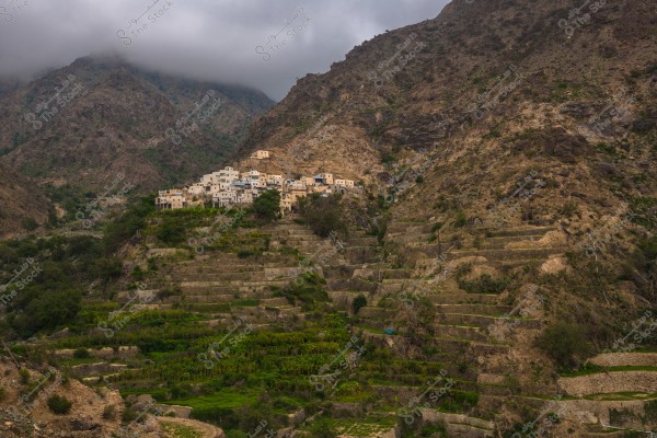 Image showing a small village nestled on terraced mountain slopes in a rugged area. The white houses are clustered on the hillside, with clouds covering some mountain peaks in the background, creating a misty atmosphere. Green plants cover the terraced slopes, indicating agricultural activity in the region.