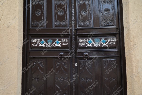 Image showing a dark-colored, intricately carved wooden gate consisting of a double door. The gate features distinctive Islamic motifs and inscriptions in Arabic, along with traditional decorative symbols. The colors used include black, blue, and white, creating a rich cultural impression. The surrounding walls appear to be old, possibly from a traditional building.