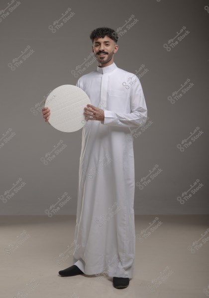 A portrait of a man wearing a traditional white thobe, standing in a studio with a grey background. The man is holding a large white circle and looking at the camera with a gentle smile.