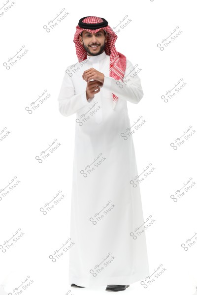 A portrait of a man wearing traditional Saudi attire, featuring a white thobe, a red ghutra, and a black agal. The man is standing with a slight smile, adjusting his sleeves, and the background is completely white.