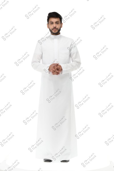 A portrait of a man standing against a white background, wearing a traditional long white garment often known as a thobe. The thobe extends to the ankles with long sleeves and a closed collar. The attire appears to reflect traditional styles found in Saudi Arabia or other Gulf countries.