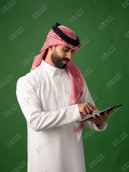صورة لرجل يرتدي ثوبًا أبيض وغترة حمراء وسوداء مع عقال, والذي يبدو وكأنه زي تقليدي خليجي, يستخدم جهازًا لوحيًا في يديه. خلفية الصورة خضراء.