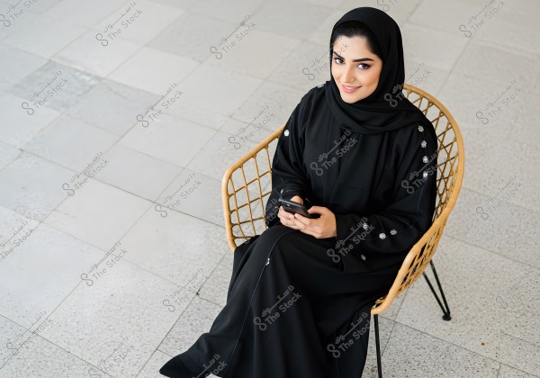 An image of a woman wearing a black abaya and hijab, sitting on a wicker chair, holding a phone in her hand and smiling at the camera. The background features light gray tile flooring, giving the photo a calm and elegant atmosphere. The woman is likely from the Gulf countries.