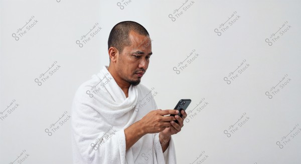 A portrait of a man wearing a white garment, likely in the context of Umrah or Hajj, using a smartphone. The background is white, giving a sense of purity and calmness.