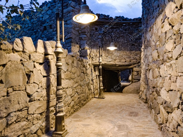 A narrow outdoor passageway lined with stone walls, illuminated by vintage street lamps. The path extends below a stone archway in the background, with a blue sky and some clouds visible above.
