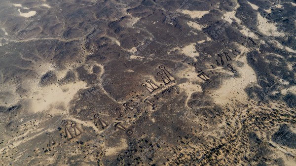 Aerial view of an archaeological site in a desert region in Saudi Arabia. The dry, expansive land features a series of geometric stone structures, including circles and rectangles, connected across the desert surface. The ancient stone formations stand out due to their dark color contrasted with the surrounding sand.