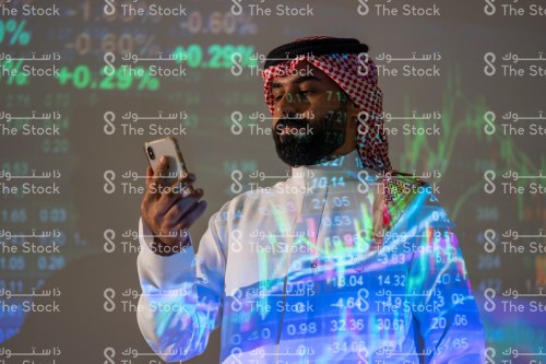 رجل سعودي يرتدي الزي السعودي التقليدي يتصفح الجوال بينما يتابع سوق الأسهم بينما تنعكس أضواء الشاشة عليه