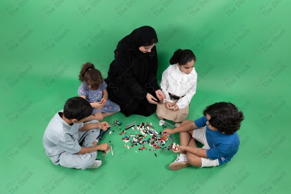 A woman in a black abaya sitting on the floor with four children "with disabilities playing with Lego blocks on a green background.