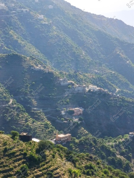 A scenic view of mountains covered with greenery and agricultural terraces. Scattered buildings, including a mosque with a minaret, are visible among the mountains. The weather appears sunny with shadows cast on the mountains.