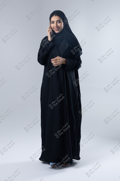 A portrait of a woman standing and smiling while talking on the phone. She is wearing a black abaya and a hijab. The background is neutral and white.