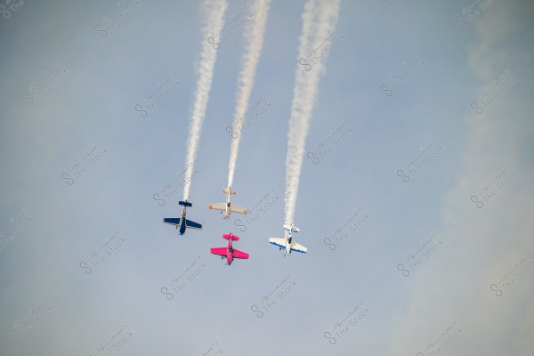 The image shows four aerobatic planes flying in a blue sky. The planes are emitting white smoke trails as they fly parallel and close to each other. The planes are colored in blue, pink, white, and orange.