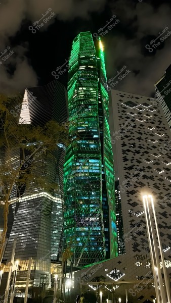An image of a skyscraper illuminated in green during a dark night. The building is surrounded by other modern towers with geometric designs and lit decorations. The ground features light poles and visible tree branches in the left corner of the image. The sky has light clouds.