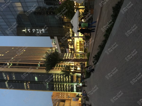 Image of a large glass building with reflected lights, next to a wide pathway with some people walking. The modern architecture and lighting suggest an urban, developed area. Palm trees and outdoor terraces under a white canopy indicate a restaurant setting.