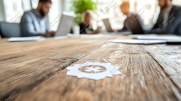 A meeting of a group of people in a modern office, focused on their laptops. A large wooden table dominates the foreground with a small white paper featuring a symbol. The surfaces are bright and organized with a green plant in the background.