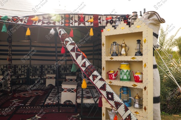 The image shows a traditional tent adorned with bright colors and geometric patterns. A wooden shelf holds traditional metal pots, including teapots and coffee pots, alongside lanterns. The tent interior is lined with traditional textiles featuring heritage designs, accompanied by traditional Arabian seating arrangements.