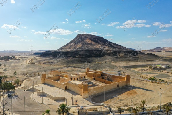 صورة تُظهر قلعة أو حصنًا تقليديًا مبنيًا من الحجارة الرملية في منطقة صحراوية مفتوحة. في الخلفية, يظهر جبل ذو قمة مسطحة. السماء صافية مع بعض السحب المتفرقة. تنتشر الأشجار والنباتات الصحراوية حول الموقع الأثري.