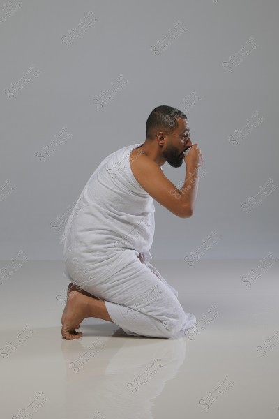 A man kneeling in a meditative position, wearing white Ihram clothing. He appears to be in an indoor setting on a reflective floor. He seems to be drinking from a small cup while wearing glasses.