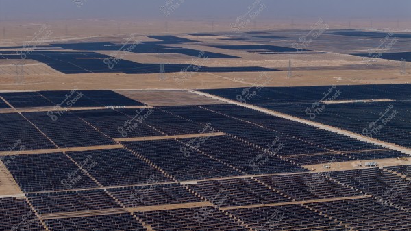 An aerial view of a massive solar farm consisting of numerous solar panels arranged in orderly rows across a vast expanse of desert land under an open sky. The panels appear dark in color, covering extensive areas, with the surrounding land having a yellowish hue.